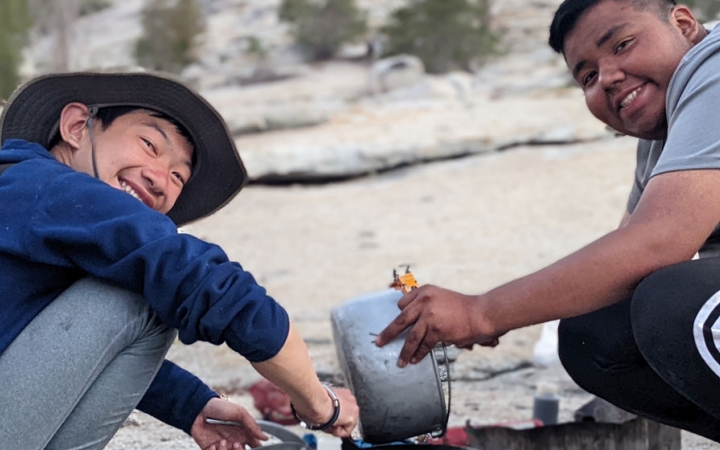 two students smile at the camera as they prepare food on a backpacking trip